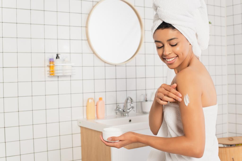 sonriente joven afroamericana mujer aplicando crema en el brazo en el baño - Foto, Imagen