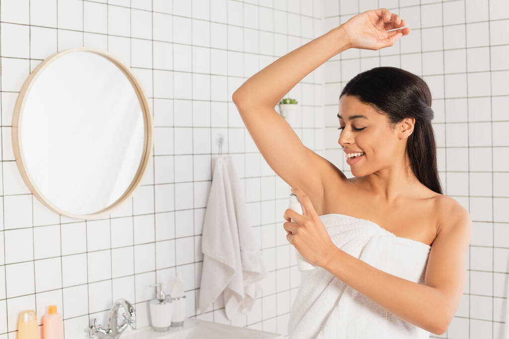 jong Afrikaans amerikaans vrouw verpakt in handdoek spuiten met deodorant in badkamer - Foto, afbeelding