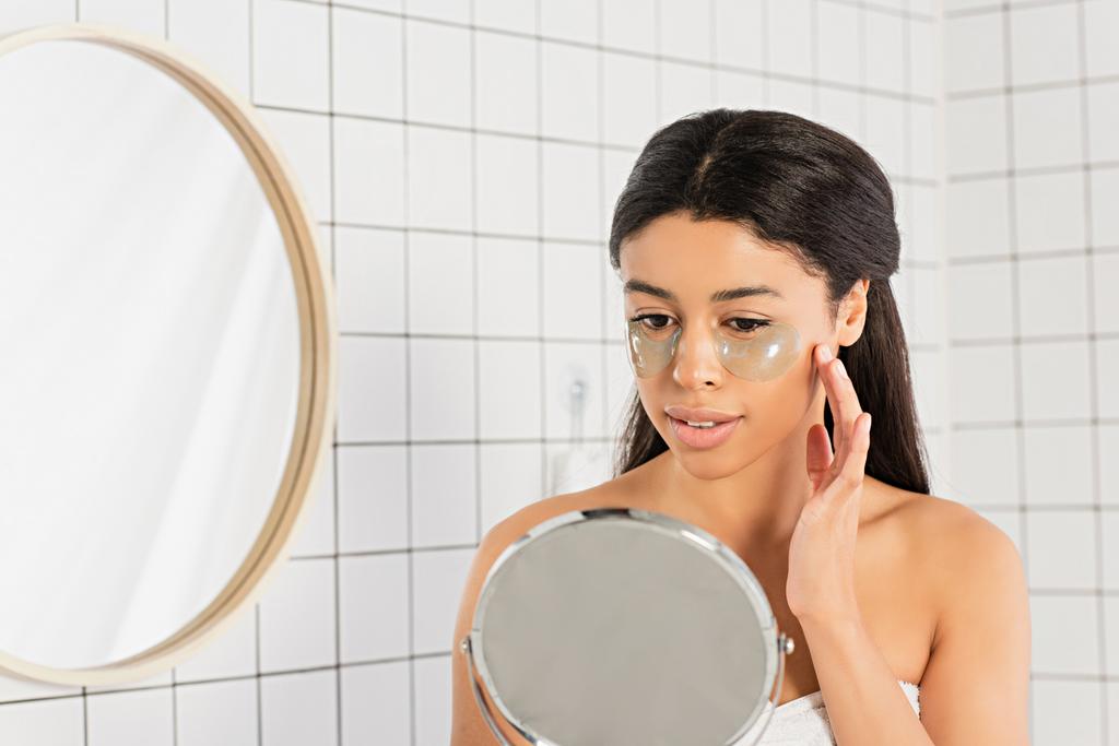 εστιασμένη νεαρή Αφροαμερικανή γυναίκα που κρατιέται χέρι-χέρι κοντά στο πρόσωπο με επιθέματα ματιών και κοιτάζει τον καθρέφτη στο μπάνιο - Φωτογραφία, εικόνα