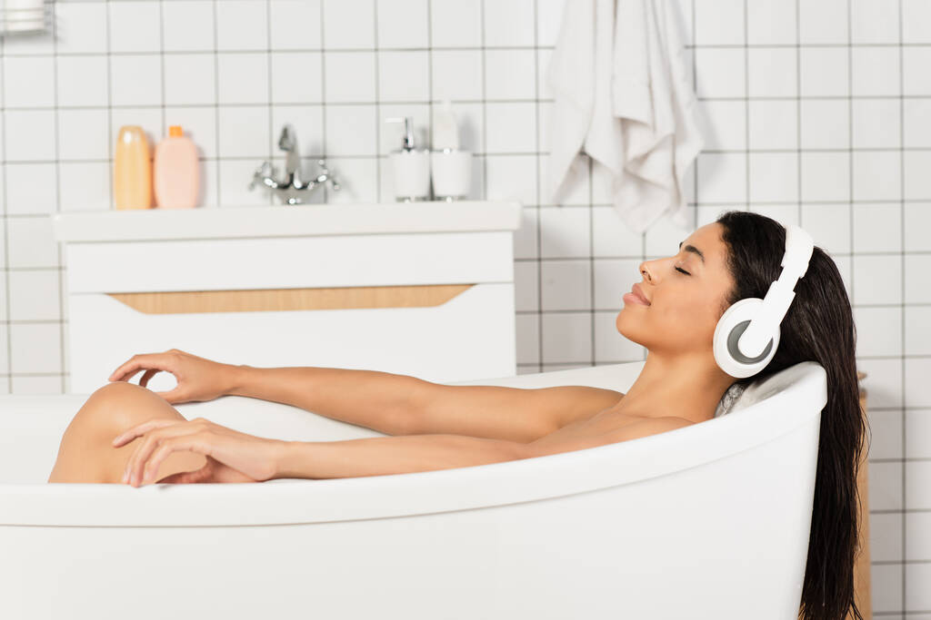 mladá žena se zavřenýma očima odpočívající a poslech hudby ve sluchátkách v koupelně - Fotografie, Obrázek