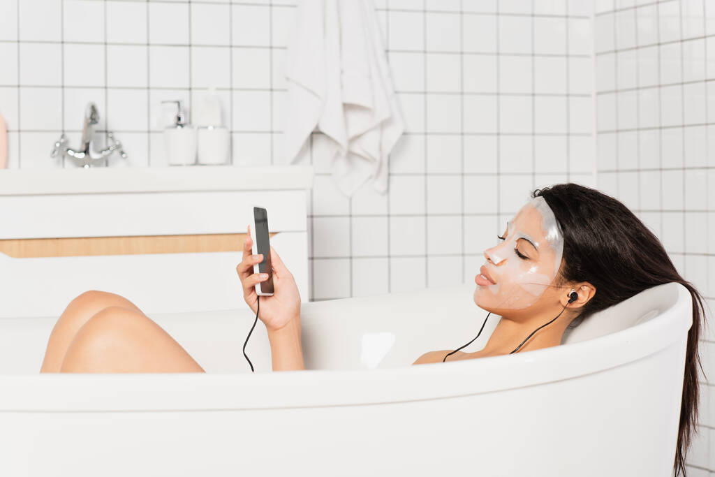 молодая женщина с маской на лице лежит с наушниками и сотовым телефоном в ванной - Фото, изображение