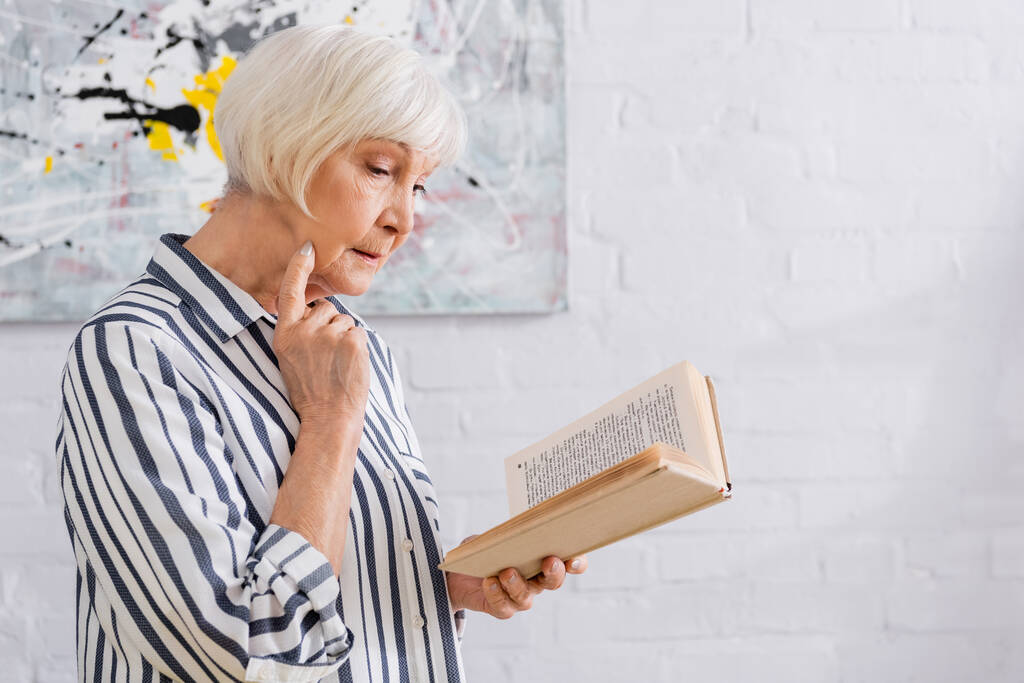 Vanhempi nainen raidallinen paita käsittelyssä kirja kotona  - Valokuva, kuva