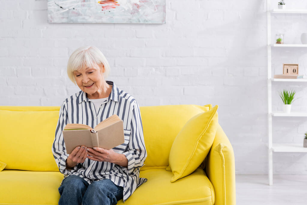 Улыбающаяся пожилая женщина читает книгу, сидя на диване  - Фото, изображение