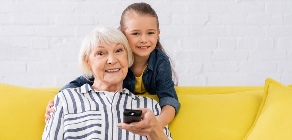 Enfant souriant étreignant grand-mère avec télécommande à la maison, bannière  - Photo, image