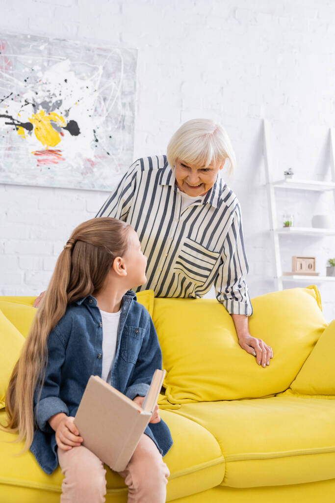 Kind met boek op zoek naar positieve oma in de woonkamer  - Foto, afbeelding