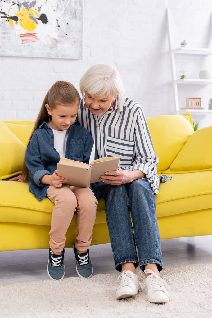 Uśmiechnięta babcia i dziecko czytają razem książkę na kanapie  - Zdjęcie, obraz