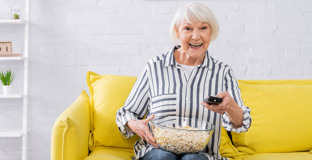 Веселая пожилая женщина с пультом дистанционного управления и попкорном дома, баннер  - Фото, изображение