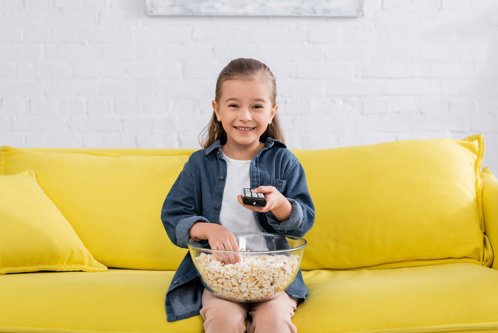 Lächelndes Mädchen mit Fernbedienung holt Popcorn aus Schüssel  - Foto, Bild