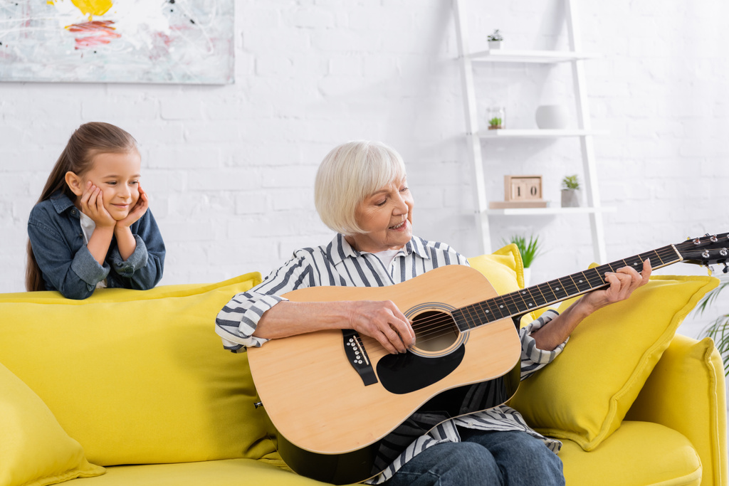 Lächelndes Kind steht neben Oma und spielt Akustikgitarre auf Couch  - Foto, Bild