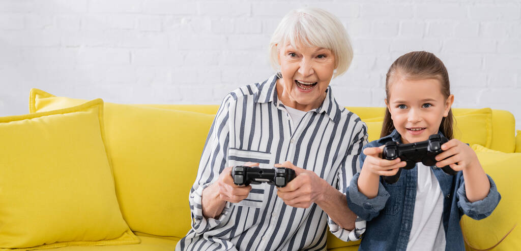 KYIV, UCRAINA - 11 GENNAIO 2021: nonna positiva che gioca ai videogiochi vicino alla nipote con joystick, striscione  - Foto, immagini