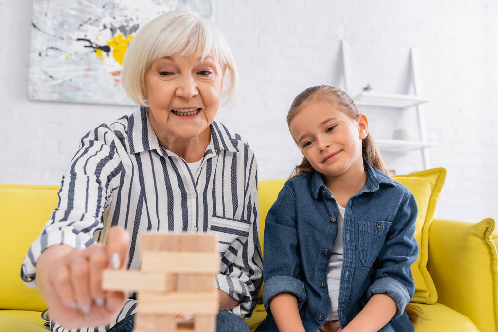 Mujer mayor sonriente jugando bloques juego de madera en primer plano borrosa cerca de la nieta en el sofá  - Foto, imagen