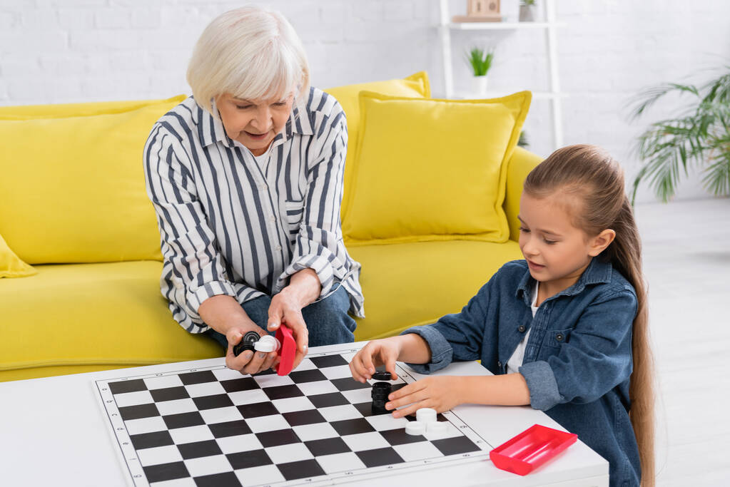 Бабушка держит шашки рядом с ребенком и журнальный столик в гостиной  - Фото, изображение