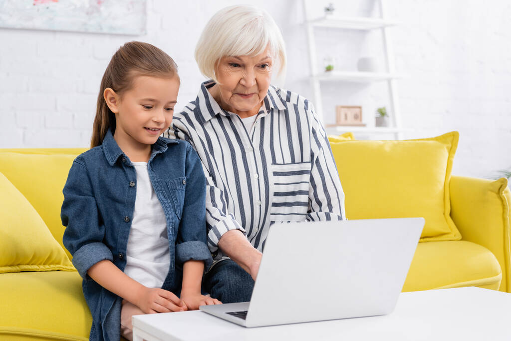 Femme âgée et enfant utilisant un ordinateur portable sur la table basse à la maison  - Photo, image