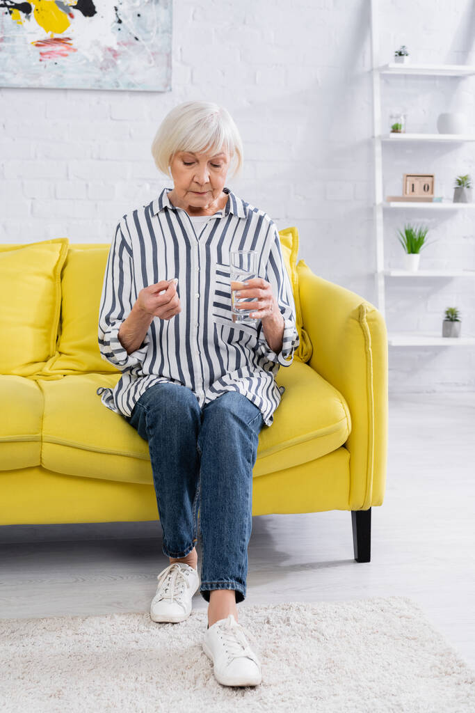 Donna anziana guardando pillola e tenendo l'acqua sul divano  - Foto, immagini