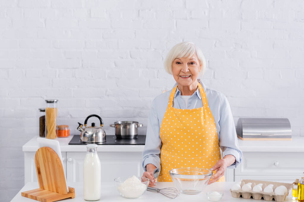 Sonriente anciana mirando a la cámara cerca de los ingredientes en la mesa de la cocina  - Foto, imagen