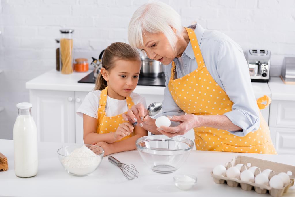 Enfant et grand-mère tenant cuillère et oeuf près du lait et de la farine dans la cuisine  - Photo, image