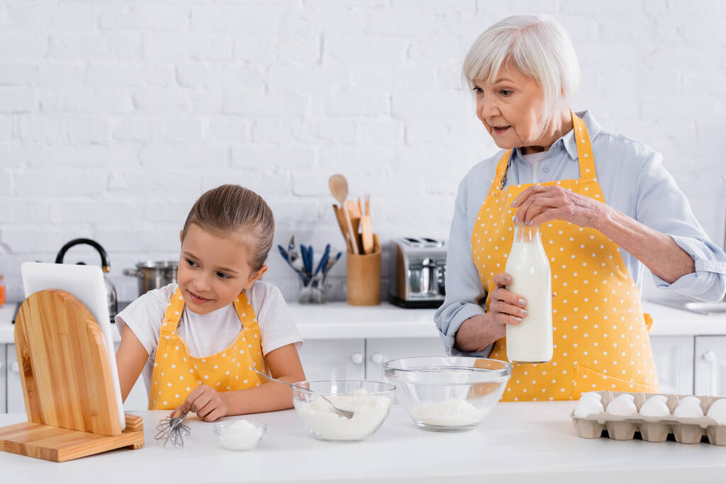 Mujer anciana sosteniendo botella de leche cerca de los ingredientes y el niño usando tableta digital en la cocina  - Foto, Imagen