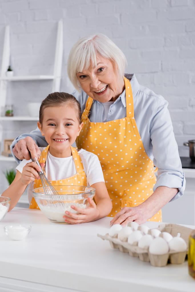 gelukkig kid en oma kijken naar camera tijdens het koken in keuken  - Foto, afbeelding