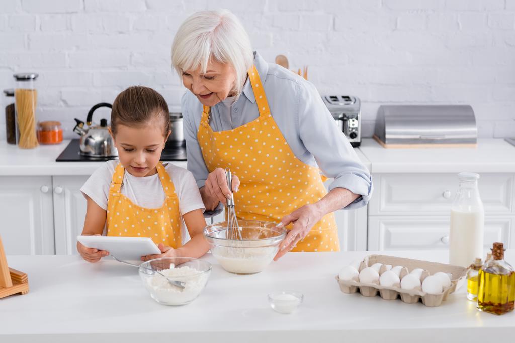 Dzieciak korzystający z tabletu cyfrowego w pobliżu babci gotowania w kuchni  - Zdjęcie, obraz
