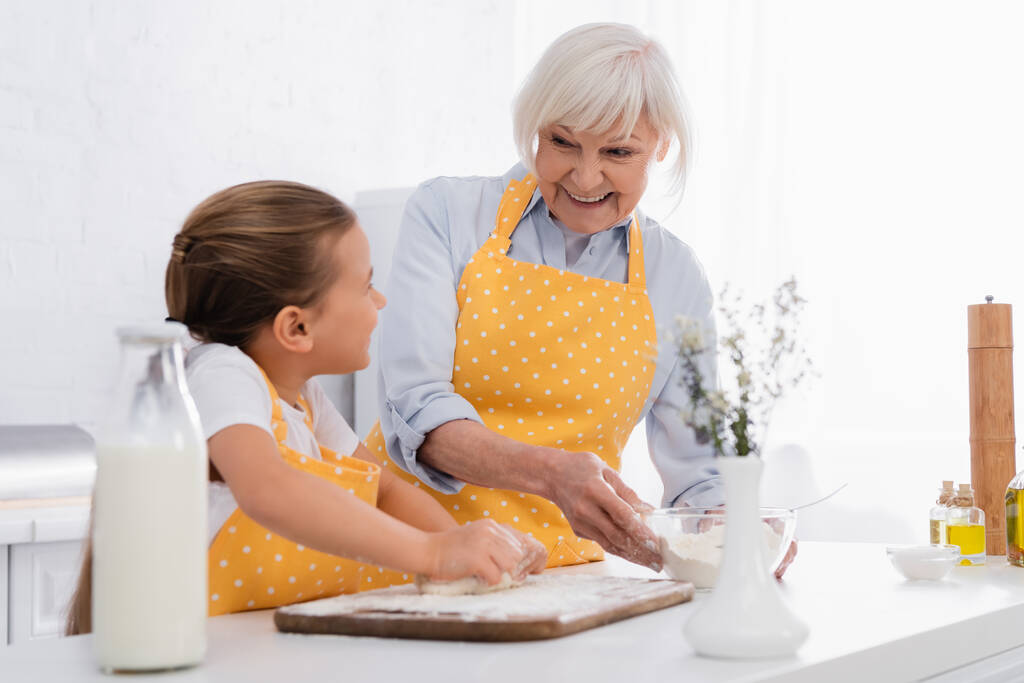 Wesoła babcia trzymając mąkę i patrząc na dziecko z ciastem na rozmytym pierwszym planie w kuchni  - Zdjęcie, obraz