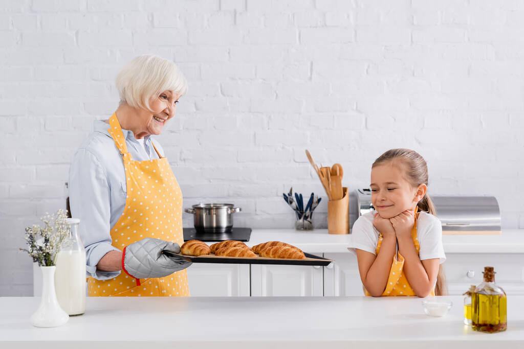 Lächelndes Kind in Schürze blickt Oma mit Croissants auf Backblech an  - Foto, Bild