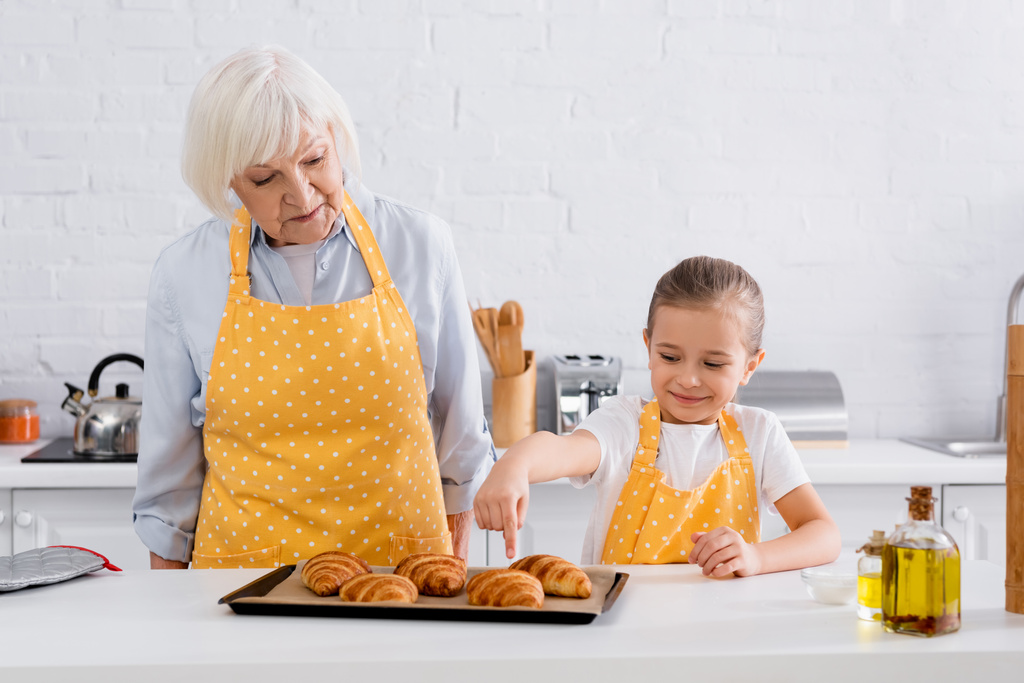 Uśmiechnięta dziewczyna w fartuchu wskazująca na rogalika w pobliżu babci w kuchni  - Zdjęcie, obraz