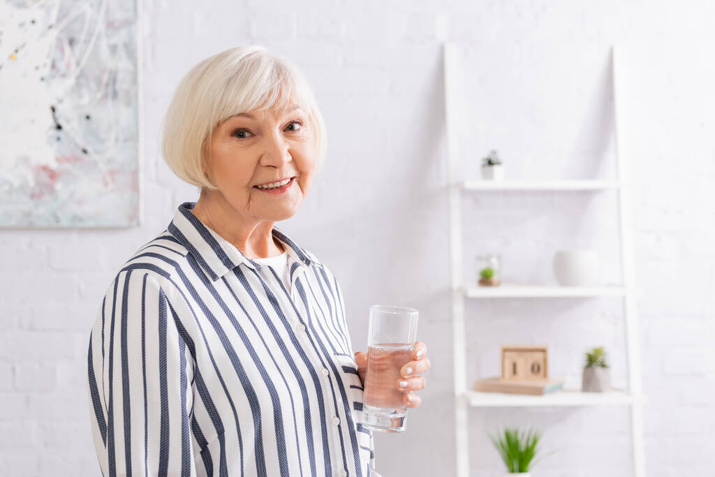 Idősebb nő egy pohár vízzel, aki otthon mosolyog a kamerába.  - Fotó, kép