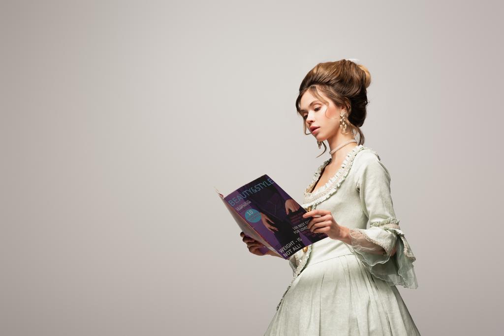encantadora mujer en vestido vintage leyendo revista moderna aislada en gris - Foto, Imagen