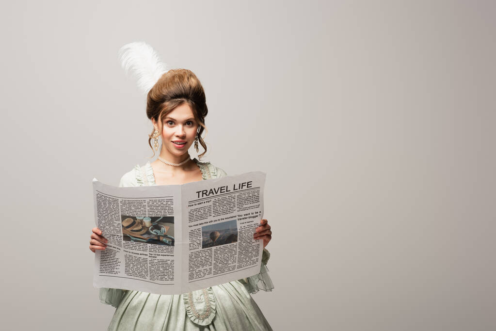 χαμογελαστή γυναίκα σε κομψό vintage στολή κρατώντας ταξιδιωτική ζωή εφημερίδα απομονωμένη σε γκρι - Φωτογραφία, εικόνα
