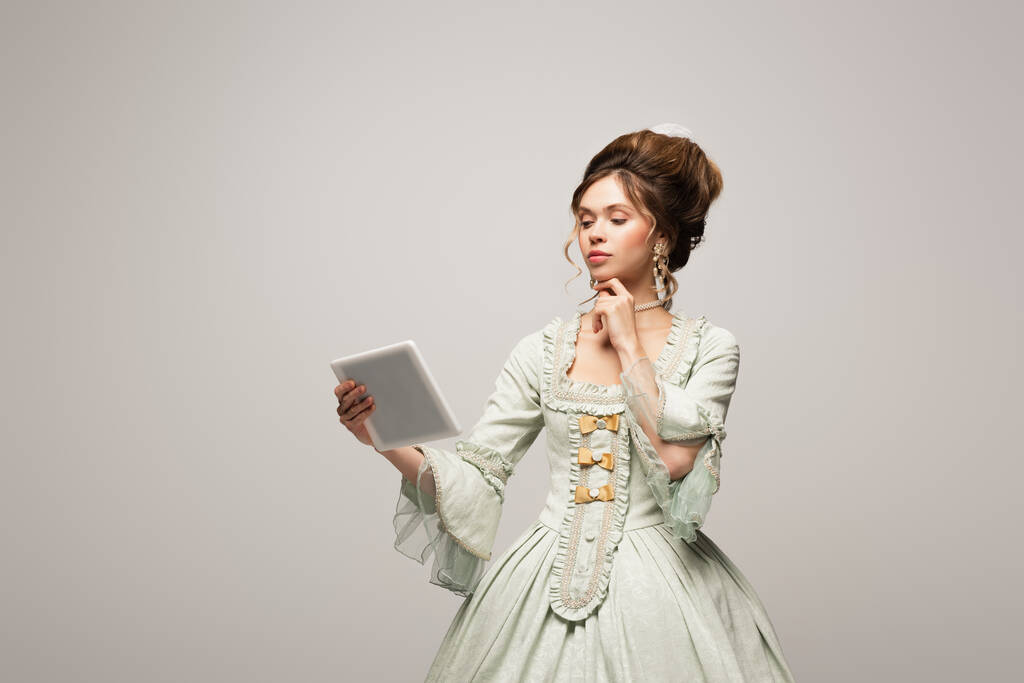 όμορφη γυναίκα σε vintage φόρεμα σκέψης, ενώ κοιτάζοντας ψηφιακή ταμπλέτα απομονώνονται σε γκρι - Φωτογραφία, εικόνα
