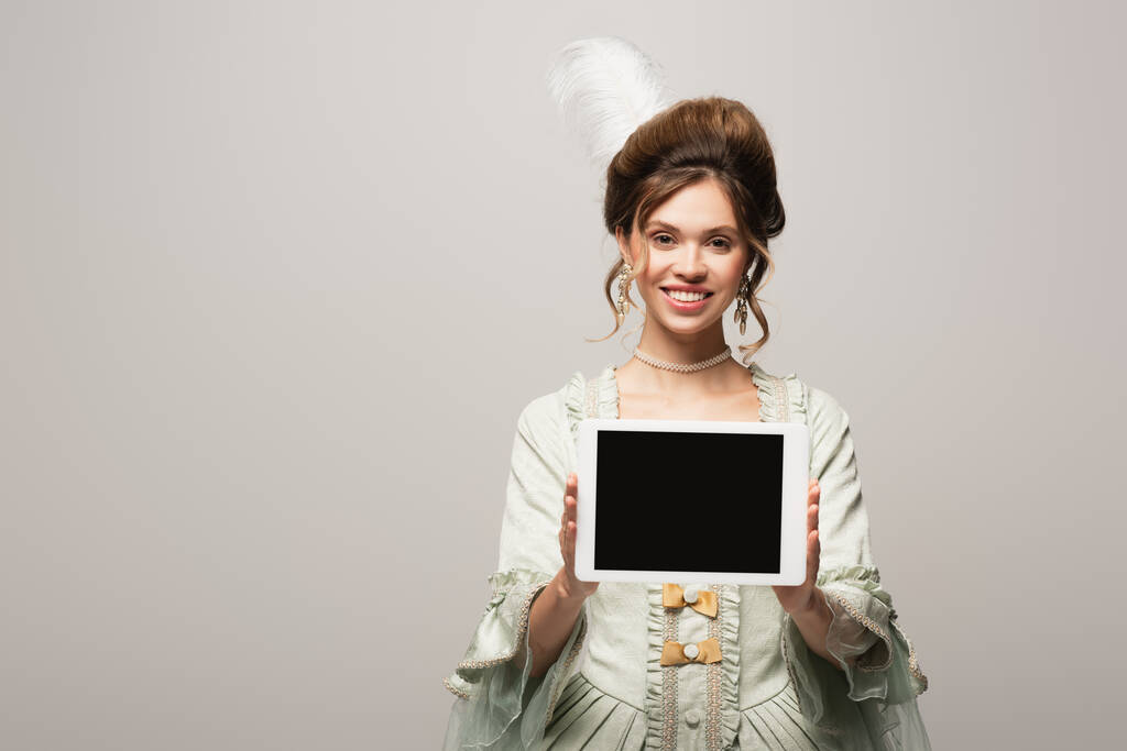 sonriente mujer de estilo retro mostrando tableta digital con pantalla en blanco aislada en gris - Foto, imagen
