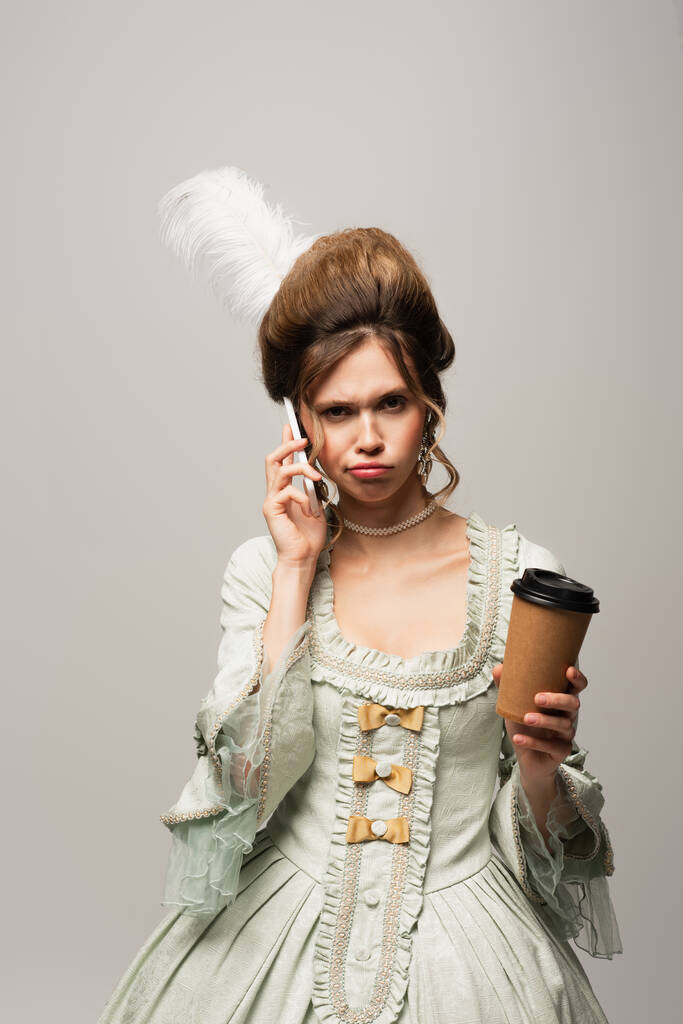 mujer disgustada en traje retro sosteniendo café para llevar mientras habla en el teléfono inteligente aislado en gris - Foto, Imagen