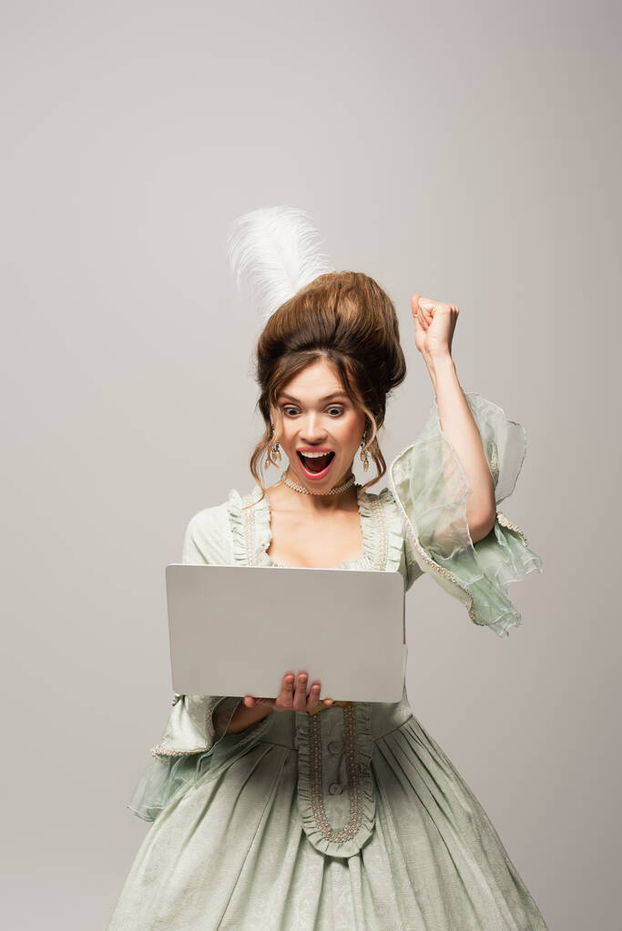 excitée femme de style rétro montrant geste de triomphe tout en regardant ordinateur portable isolé sur gris - Photo, image