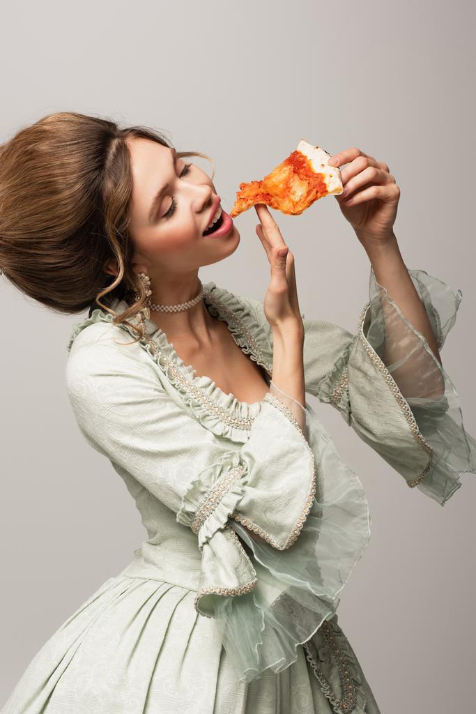 mujer joven en elegante vestido vintage comiendo pizza con los ojos cerrados aislados en gris - Foto, Imagen