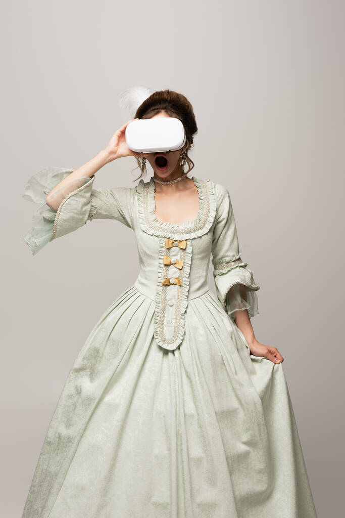 взволнованная женщина в ретро платье касаясь VR гарнитуры в то время как игры изолированы на серый - Фото, изображение