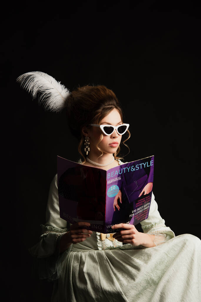 стильная женщина в винтажном наряде и в модных солнцезащитных очках читает современный журнал красоты и стиля, изолированный от черного - Фото, изображение