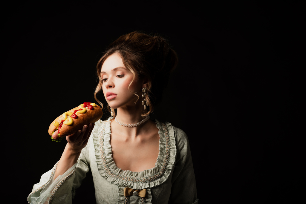 pretty woman in elegant retro dress holding delicious hot dog isolated on black - Photo, Image