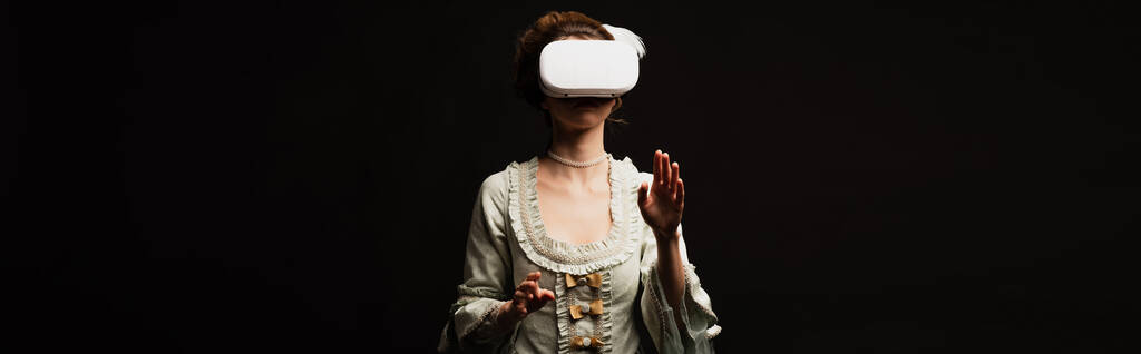 молодая женщина в платье в стиле ретро жесты в VR гарнитуре изолированы на черном, баннер - Фото, изображение