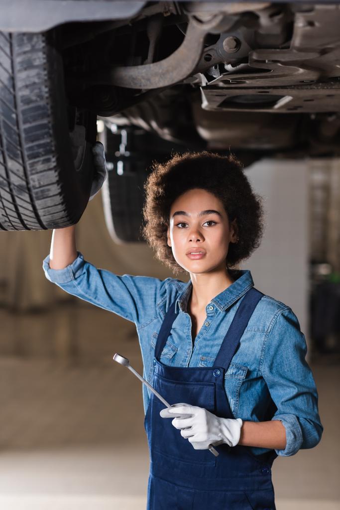 joven afroamericano mecánico celebración de la llave en la mano y de pie debajo del coche en el garaje - Foto, Imagen
