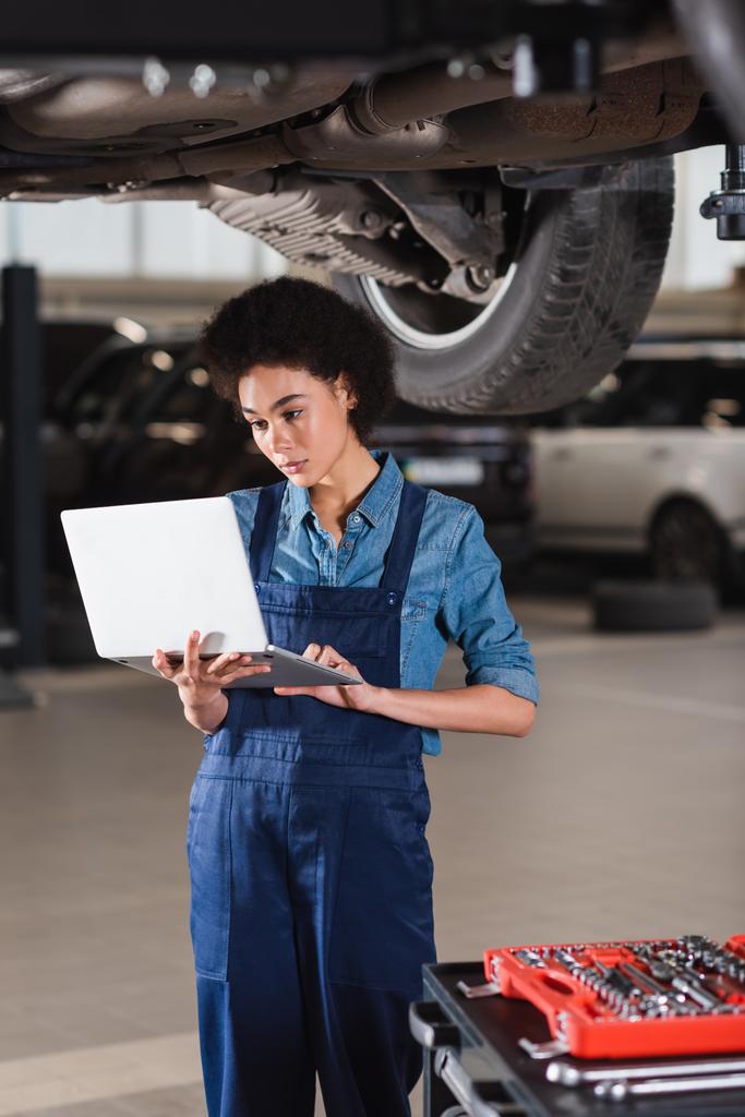 young african american mechanic standing underneath car and typing on laptop in garage - Photo, Image