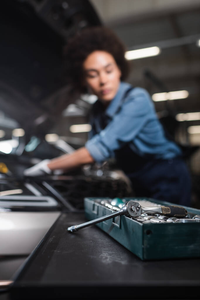 Nahaufnahme von Schraubenschlüssel in Werkzeugkiste mit verschwommenem afrikanisch-amerikanischem Mechaniker bei der Arbeit mit Auto in Garage - Foto, Bild