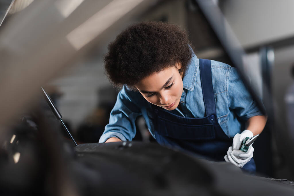 giovane meccanico afroamericano in tuta con pinze e che lavora con auto in garage  - Foto, immagini