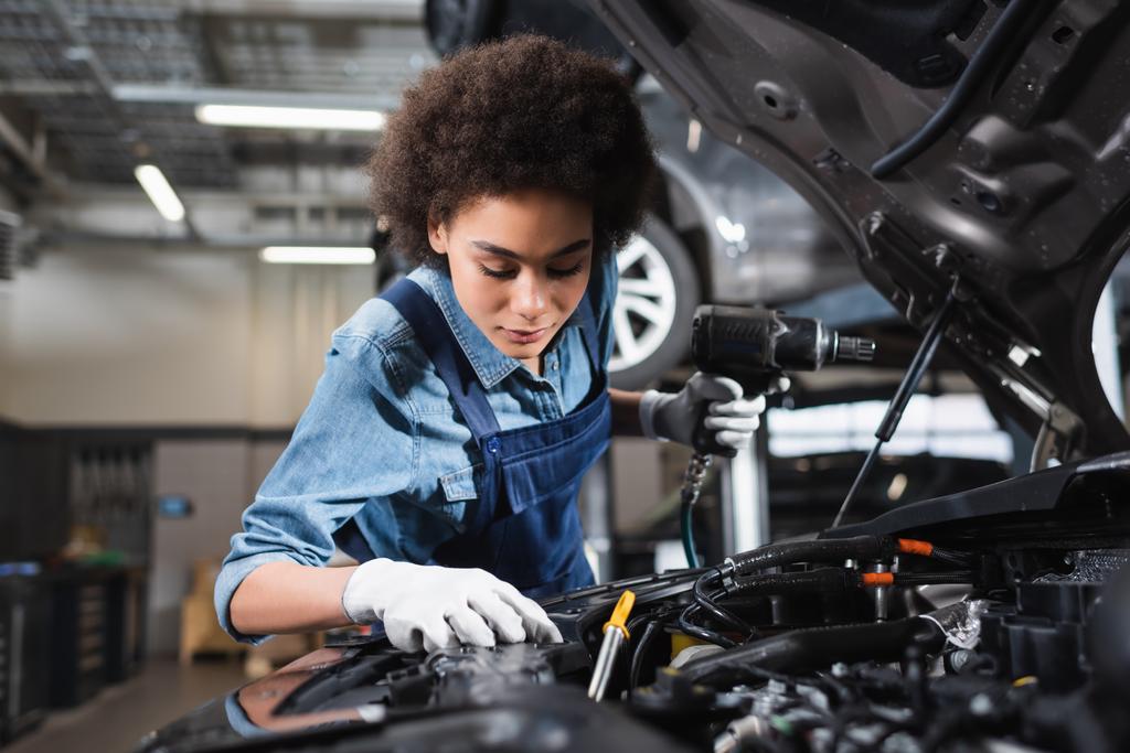 mladý africký americký mechanik v montérkách drží elektrický šroubovák a pracuje s motorem v garáži - Fotografie, Obrázek