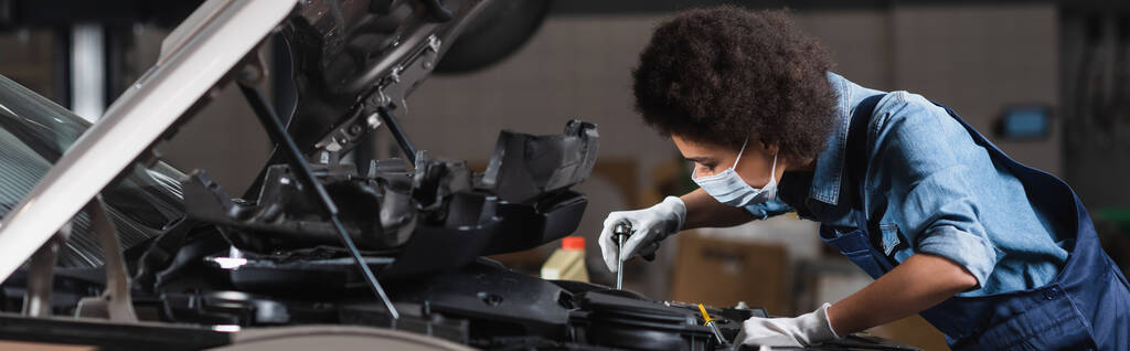 joven mecánico afroamericano en máscara protectora que sostiene la llave inglesa y que trabaja con el motor del coche en el garaje, bandera - Foto, imagen