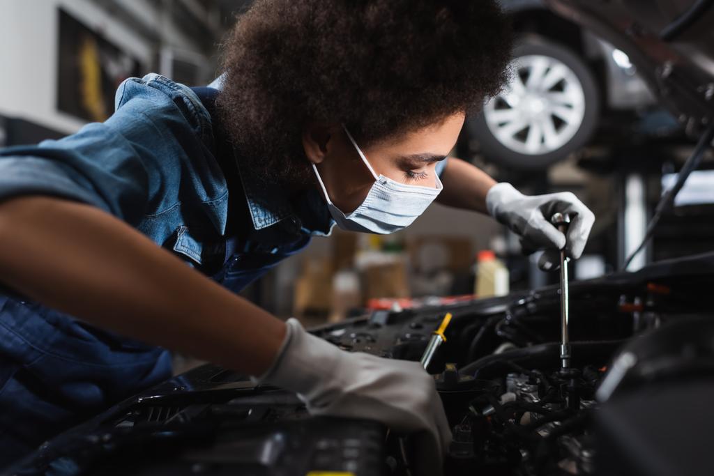 jovem afro-americano mecânico em máscara protetora trabalhando com motor de carro na garagem - Foto, Imagem