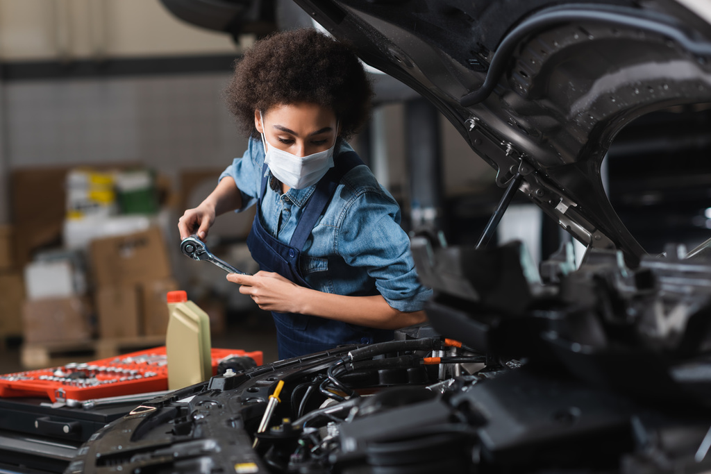 joven afroamericano mecánico en máscara protectora trabajando con motor de coche en garaje - Foto, Imagen