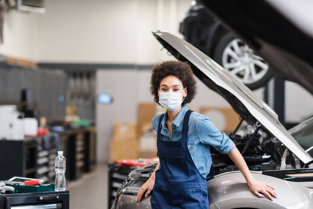 jovem afro-americano mecânico em máscara protetora de pé perto do carro na garagem - Foto, Imagem