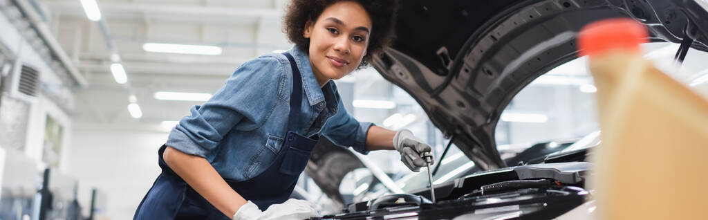 sonriente joven afroamericano mecánico sosteniendo la llave y trabajando con el motor del coche en el garaje, pancarta - Foto, Imagen