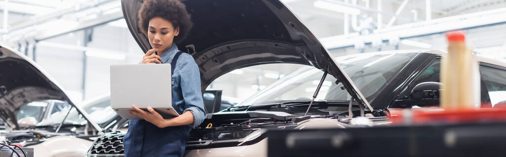serious young african american mechanic holding laptop near car with open hood in auto repair service, banner - Photo, Image