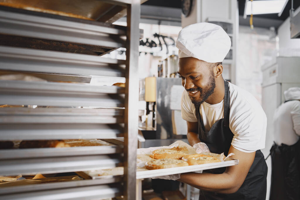 Afroamerikanischer Bäcker in Uniform sortiert Backwaren auf Paletten - Foto, Bild